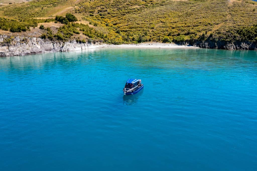 Aerial view of fishing charter boat in clear bay at Lake Hāwea with beach and mountains