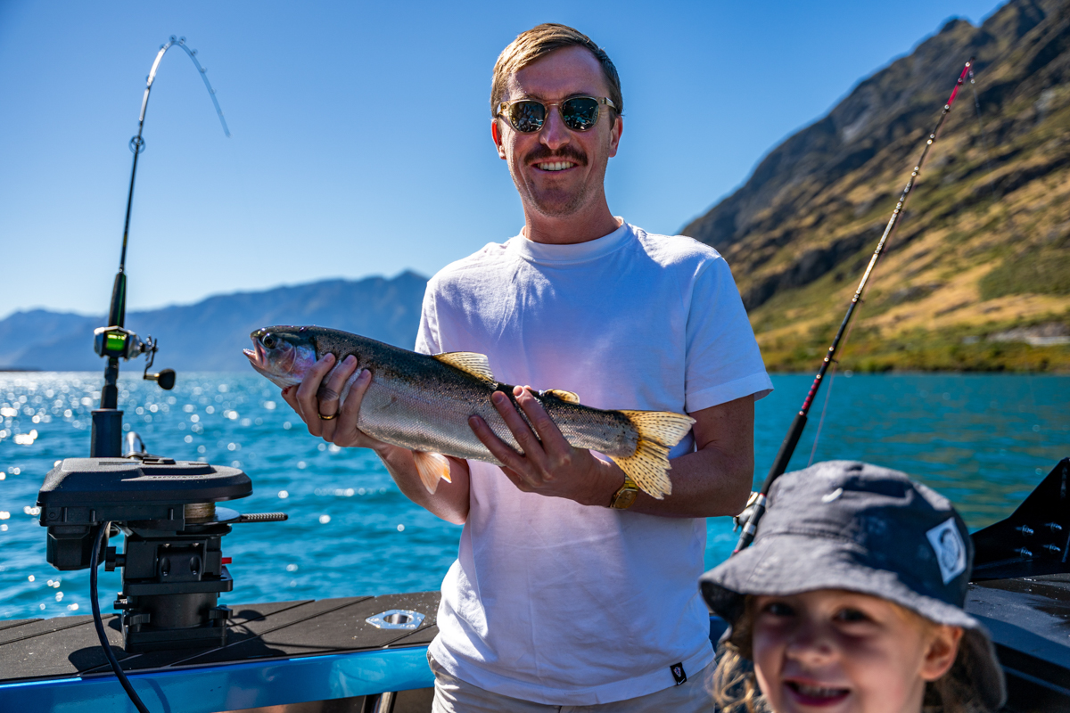 Man proudly holding a trout on a fishing boat at Lake Hāwea, New Zealand