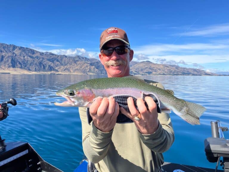 Hunter smiling with a rainbow trout catch on a beautiful day at Lake Hāwea