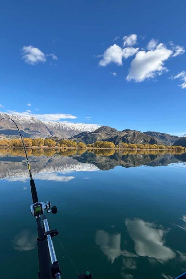 flat-lake-mountains-fishing-rod-nz-charter Flat lake surface with mountain backdrop and fishing rod in the foreground during a New Zealand charter